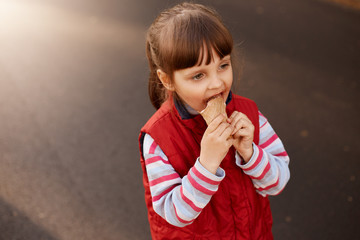 Close up portrait of funny playful little kid being outside, having walk, looking ahead, holding food in hands, eating ice cream, wearing casual clothes, enjoying vacation. Childhood concept.