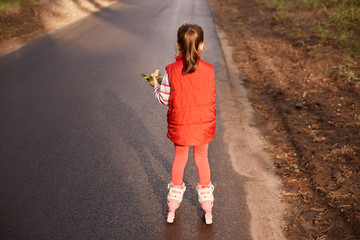 Back view of tiny little girl with ponytail wearing red casual clothes, holding food in one hand,...