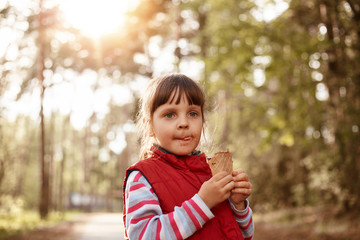 Portrait of pretty playful obedient kid looking ahead, holding ice cream in hands, playing outside, posing over trees background, being in park, having thoughtful facial expression. Rest concept.