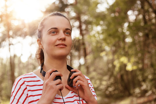 Horizontal shot of dreamy good looking attractive young lady having walk, being in unity with nature, having headphones around neck, breathing fresh air, wearing striped sweatshirt. Nature concept.