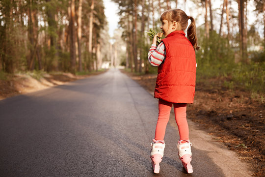 Picture of joyful active little girl eating ice cream, rollerskating alone, wearing red suit, having ponytail, being near forest, having fun, enjoying holidays, being fond of nature. Rest concept.