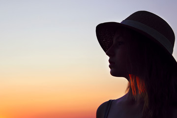Blurry silhouette of teenage girl wearing a sun hat and looking to the left, close up portait, space for text. Female portrait, sunset background, cropped shot. 
People, travel concept.