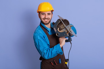 Horizontal photo of good looking handsome happy worker holding tool in both hands, looking directly at camera, smiling sincerely, wearing brown apron, blue overall and yellow helmet. Work concept.