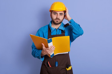 Portrait of emotional magnetic helpless young builder looking directly at camera, holding papercase, putting one hand on helmet, having working day, being busy. People and work problems concept.