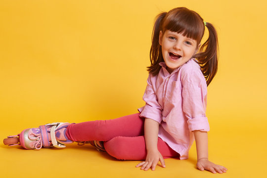 Horizontal picture of little girl opening mouth widely, being on floor, looking directly at camera, wearing roller blades, being in good mood, having fun. Children and leisure activities concept.