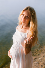 Fototapeta premium Pregnant blonde woman in long white dress on the beach. Future mother with belly