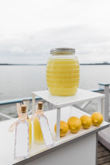 Four yellow lemons and a carafe with lemon juice on a white table outside. limoncello