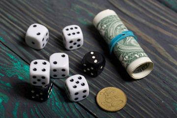 Dices are white and black. Nearby dollars rolled up into a tube and a coin. On brushed pine boards.
