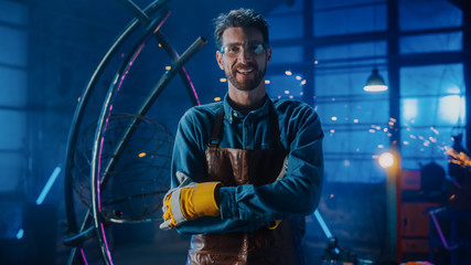 Young Professional Fabricator in Safety Glasses and Apron Gently Smiles at the Camera with Crossed Arms. Authentic Artist Wearing Work Clothes in a Metal Workshop. Sparks Flying on the Background.