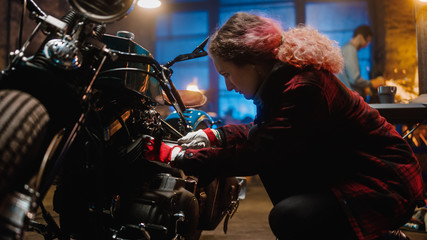 Obraz na płótnie Canvas Young Beautiful Female Mechanic is Fixing a Custom Bobber Motorcycle. Talented Girl Wearing a Checkered Shirt. She Uses a Ratchet Spanner. Creative Authentic Workshop Garage.