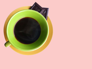 Green cup of black coffee on a yellow plate with dark chocolate on a pink background. View from above.