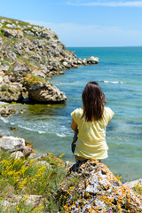 a girl sits with her back to us on a rock on a rocky coast in Sunny weather and looks out to sea