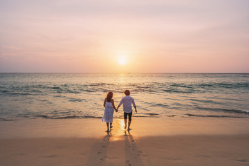 Young woman traveler holding man's hand and looking beautiful sunset on the beach, Couple on vacation in summer concept