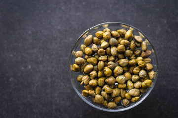 pickled capers in a glass bowl on a dark background, top view close up