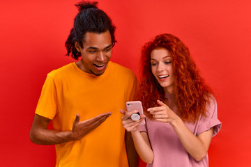 Young mixed race couple african american man and charming caucasian woman watching online store with furniture using smartphone and Internet on red background. Relocation concept. Advertising space