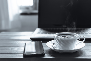 A white cup of coffee with smartphone on the desk, Morning coffee time with notebook computer and work equipment, Normal practice by working at home during covid-19 outbreak around the world.