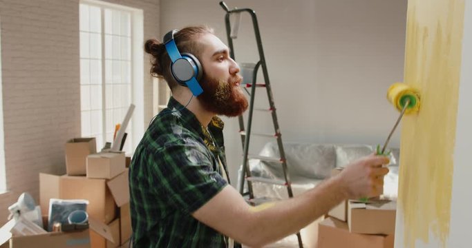 Bearded hipster caucasian guy moved to a new apartment. Young man renovating his house, painting the wall while listening to music - new life concept close up 4k footage