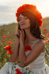 Young beautiful woman with closed eyes. In a white dress and a wreath of poppies on her head, she sits in a field of poppies at sunset with her hands folded under her chin.