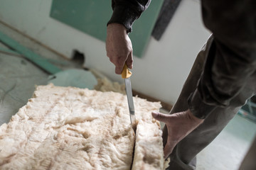 Handwerker schneidet mit einem großen Messer Isolierung aus Glaswolle passend zu auf einer...