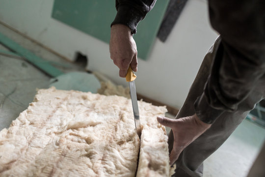 Handwerker schneidet mit einem großen Messer Isolierung aus Glaswolle passend zu auf einer Baustelle im Eigenheim