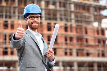 Happy young Caucasian civil engineer or businessman standing in front of a construction site and showing a thumb up, holding rolled building plans under his arm