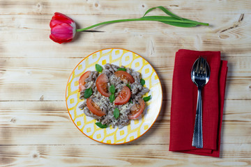 brown rye pasta with white sauce, basil leaves, tomato, cheese, pepper in  plate on wooden background with red tulip and cutlery