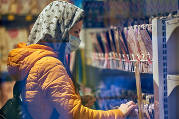 A girl in a medical mask, hood and briefcase, chooses an item in the store. Lady in an orange jacket. was tired of covid-19. the background is indistinct. counter