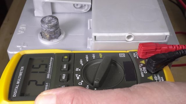 A man&rsquo;s hands switching on a digital multimeter next to a vehicle battery, then taking a reading by connecting the red probe to the positive terminal, to check the battery is  charged.