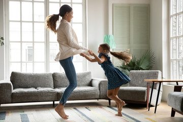 Overjoyed young mom or nanny have fun jumping in living room with excited small girl child, happy...
