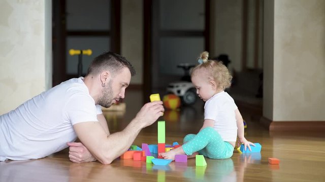 Father is playing with a little daughter. Dad and baby have a great time building a tower of colored figures on the floor