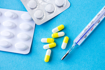 analog thermometer on blue background with bunch of medicaments in white/yellow capsules