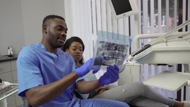 Male african dentist speaking with patient, pretty dark skinned girl, in dental clinic, showing her panoramic dental X-ray of the jaws. Dentistry and oral care concept