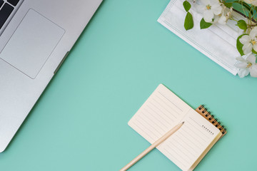 notebook with a pencil, a laptop, a mask and a blossoming branch of an apple tree on the desktop. Work at home during the quarantine period. Mint color background