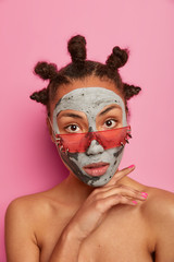 Vertical shot of serious calm woman stands with naked body, keeps hand under chin, applies clay mask, cares about skin, wears stylish sunglasses, looks beautiful. Face care and spa treatments