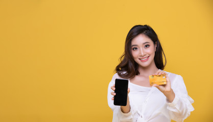 Portrait Asian beautiful happy young woman smiling cheerful and she is holding credit card and using smart phone for shopping online on yellow background.