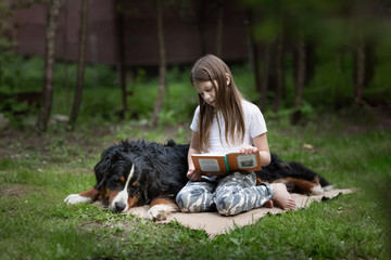 Caucasian little girl child reads a book to a large dog in a summer glade, a child with a Bernese Mountain Dog pet, friendship of a child and a dog, Companion dog and dog therapy concept.