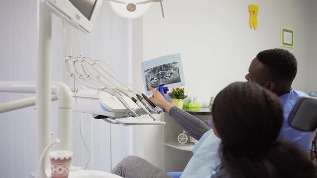 Side angle view of female african lady patient, speaking with her doctor dentist, handsome African man in blue medical suit, in modern dental clinic, looking at xray image of the jaws.