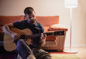 Obraz premium Young man sitting on the floor and leaning the back on the sofa playing the guitar at night time with the light of a lamp