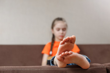 A little caucasian girl with bare feet reading book while sitting on a sofa at home. Foot closeup....