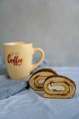 Two sponge cake roll slices are laying on wooden cutting board against background of a cup of morning coffee