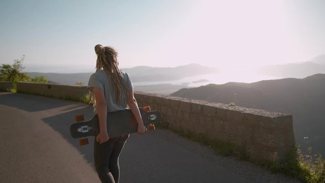 Camera following dreadlocks woman walking with longboard on mountain background cruising downhill on road а enjoying amazing view summer vacation, Montenegro
