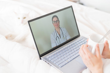 A faceless woman drinks an antipyretic and watches a video from a doctor recommendation. Online doctor consultation on a webcam without leaving your home. The cat lies in bed with a sick housewife.