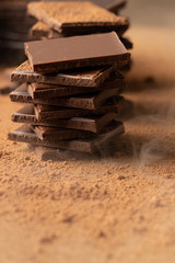 Pieces of dark chocolate bars against a background of cocoa powder and light smoke. Vertical format