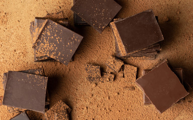 Folded and broken bars of dark chocolate on a brown background sprinkled with cocoa powder, top view