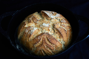 Freshly baked round bread loaf with scoring designs and patterns inside a cast-Iron skillet on black dark background, homemade gluten-free artisan bread loaf 