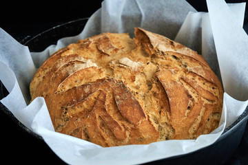Close up freshly baked round bread loaf with scoring designs and patterns inside a cast-Iron skillet on black dark background, homemade gluten-free artisan bread loaf