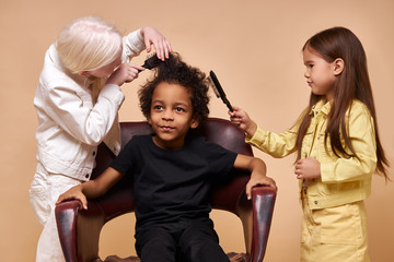 albino child girl combing curly hair of africanamerican boy isolated, girl brushing hair of cute boy sitting on chair. caucasian girl with long help albino girl