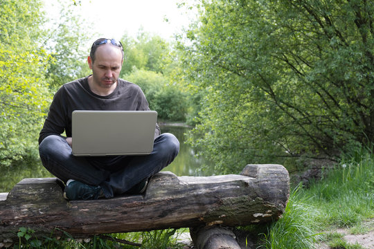 a man with a laptop sits on a log near the lake