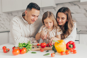 Happy family of little girl and her parents having fun and cooking salad in kitchen at home.