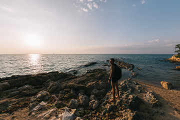 A man with a backpack stands on a rocky shore against the background of the sea and sunset sky